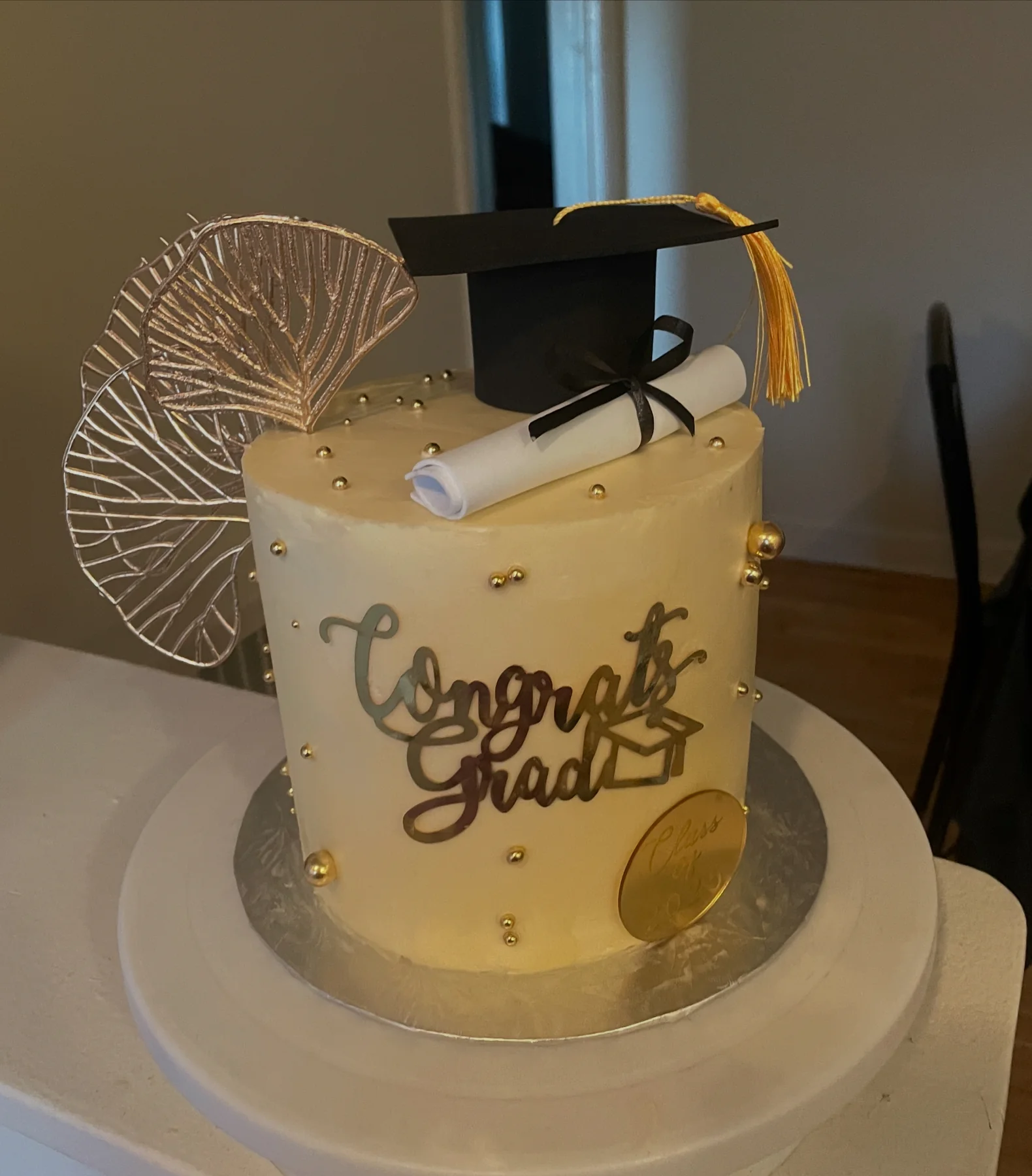 Cream graduation cake decorated with gold fans, a cap, and a diploma