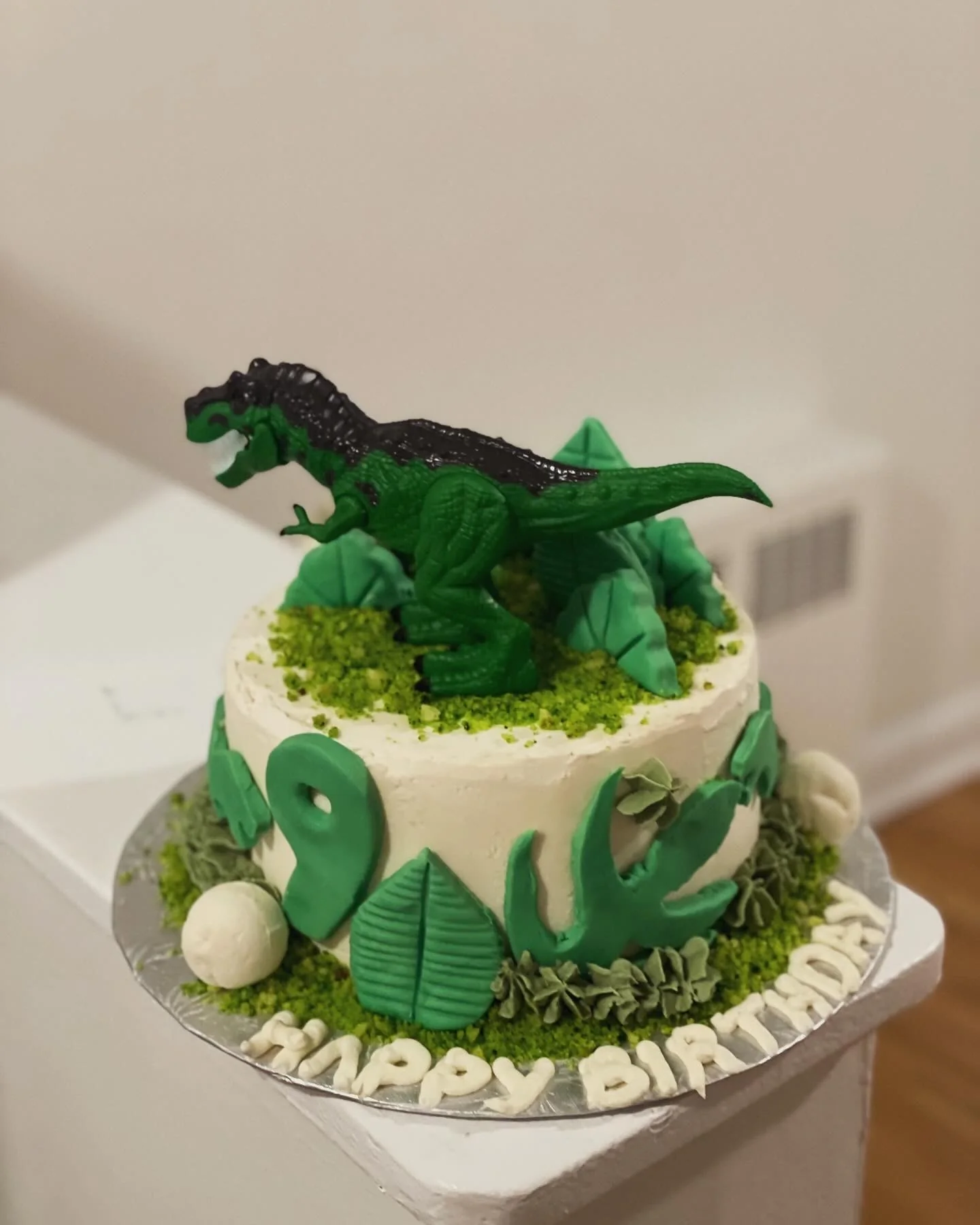 Green dinosaur birthday cake with leaves, rocks, and a toy dinosaur topper